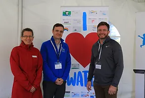 a woman and two men, wearing business clothes stand in front of an 'I heart water' sign and smile