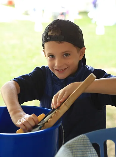 a young boy wearing a backwards baseball cap and blue shirt plays in a bucket of water and smiles at the camera