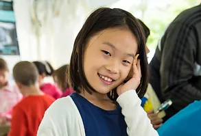 a young girl with short black hair and a navy blue shirt holds her hand to her face and smiles for a photo