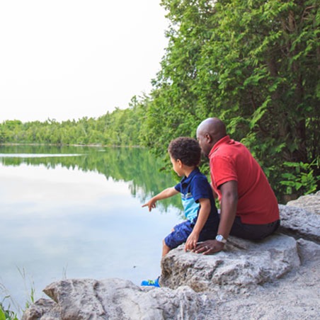 An adult and their child sit on rocks at the edge of the lake. The child points at something across the lake.