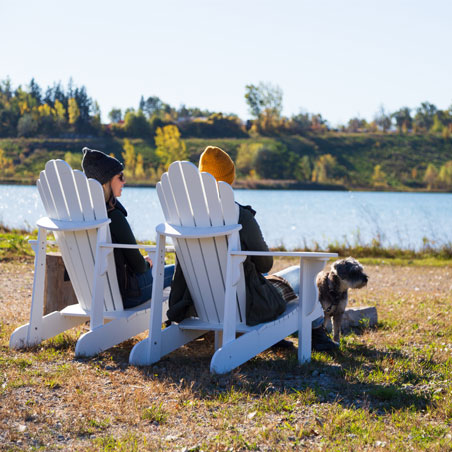 Two people sit by the lake in white Muskoka-style chairs with their dog on a leash, during early fall.