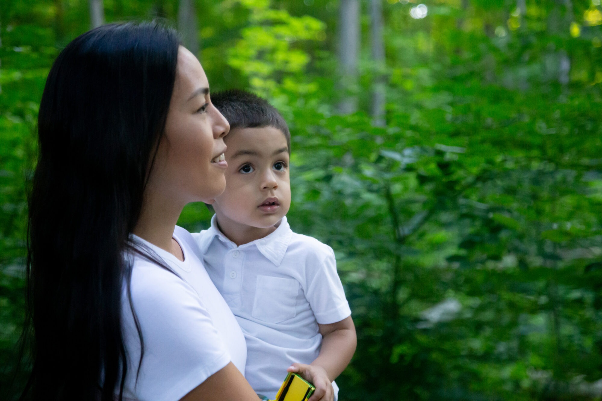 a woman holds her son, both look off camera. green trees and bushes can be seen behind them