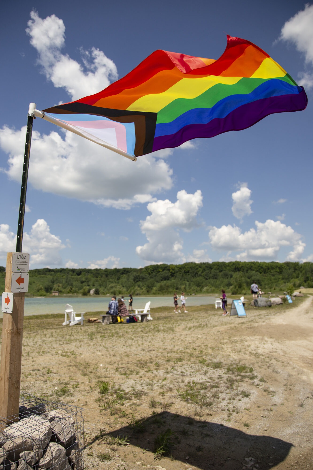 a pride flag in the wind, a beach with people sitting in the sand can be seen in the background